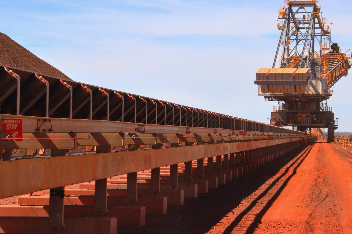 A large mining conveyor in an open-pit mine transports materials. The machinery stretches along a red dirt landscape under a blue sky.