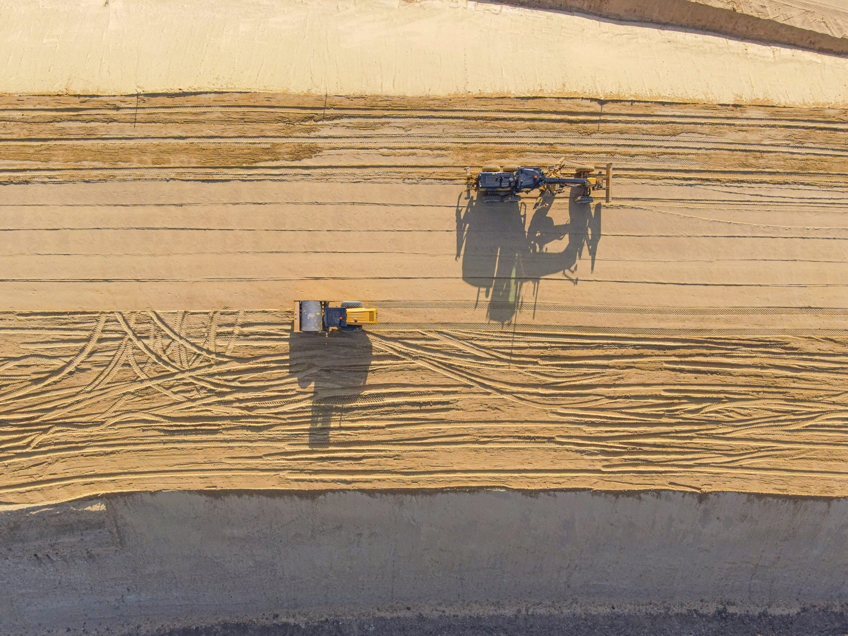 Construction roller and grader on sand, aerial view Construction roller and grader on sand, aerial view - photo of construction equipment on a sand road. Construction roller on the sand. A grader on the sand. Bulldozer on the sand. A sandy road. Shadow of construction equipment on the sand. Photo from a drone.